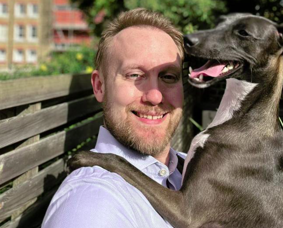 A man with a beard smiles while holding a happy dog outside on a sunny day. They are seated on a wooden bench, with trees and a brick building in the background.